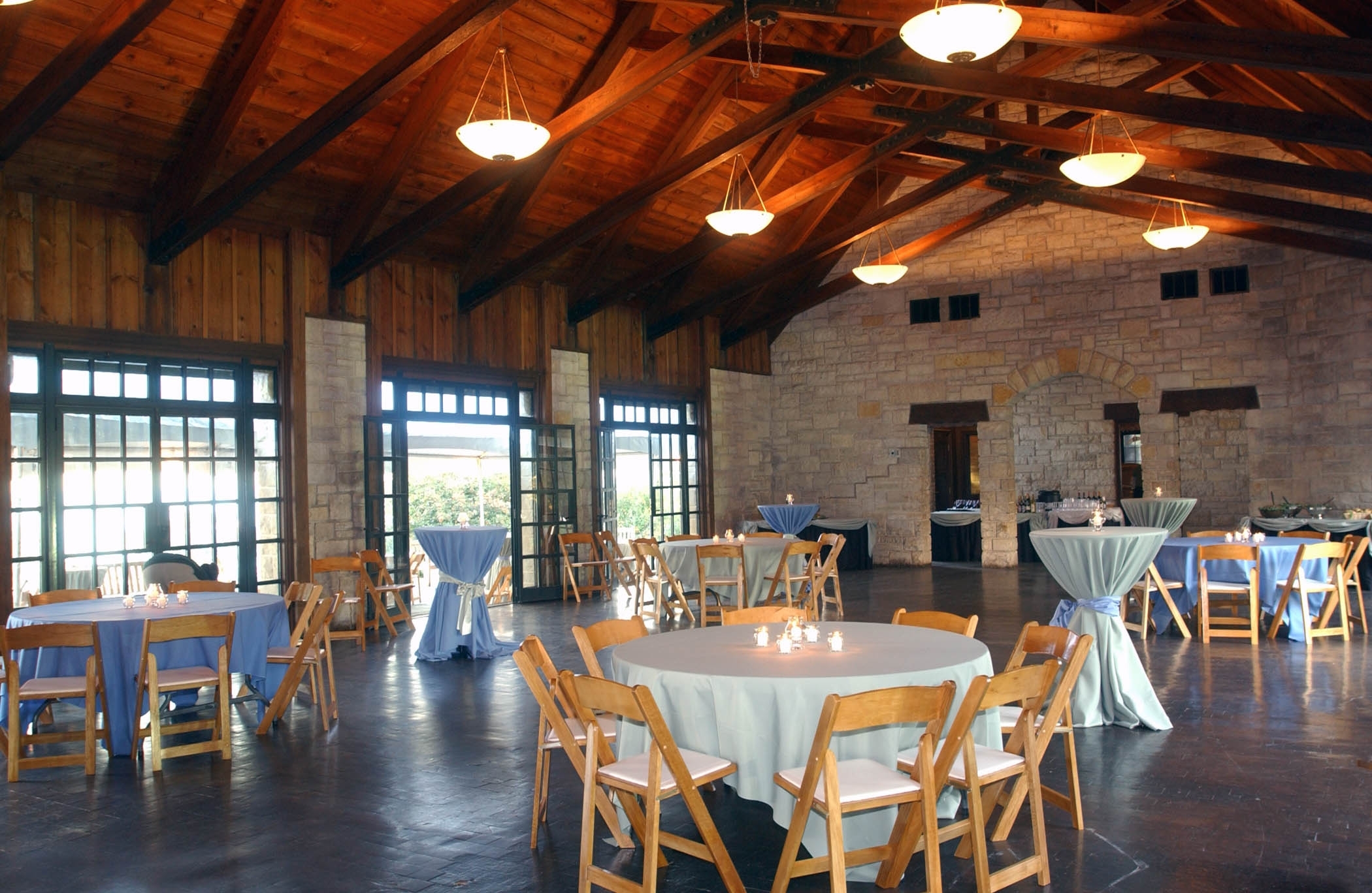 Rustic event hall with round tables, wooden chairs, and light blue tablecloths set up for a reception. Wood beam ceiling and stone walls.